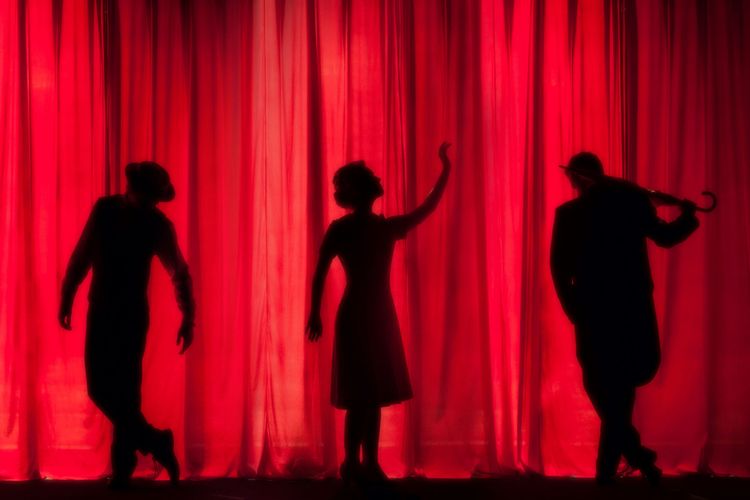 Three silhouetted figures set against a red curtain 